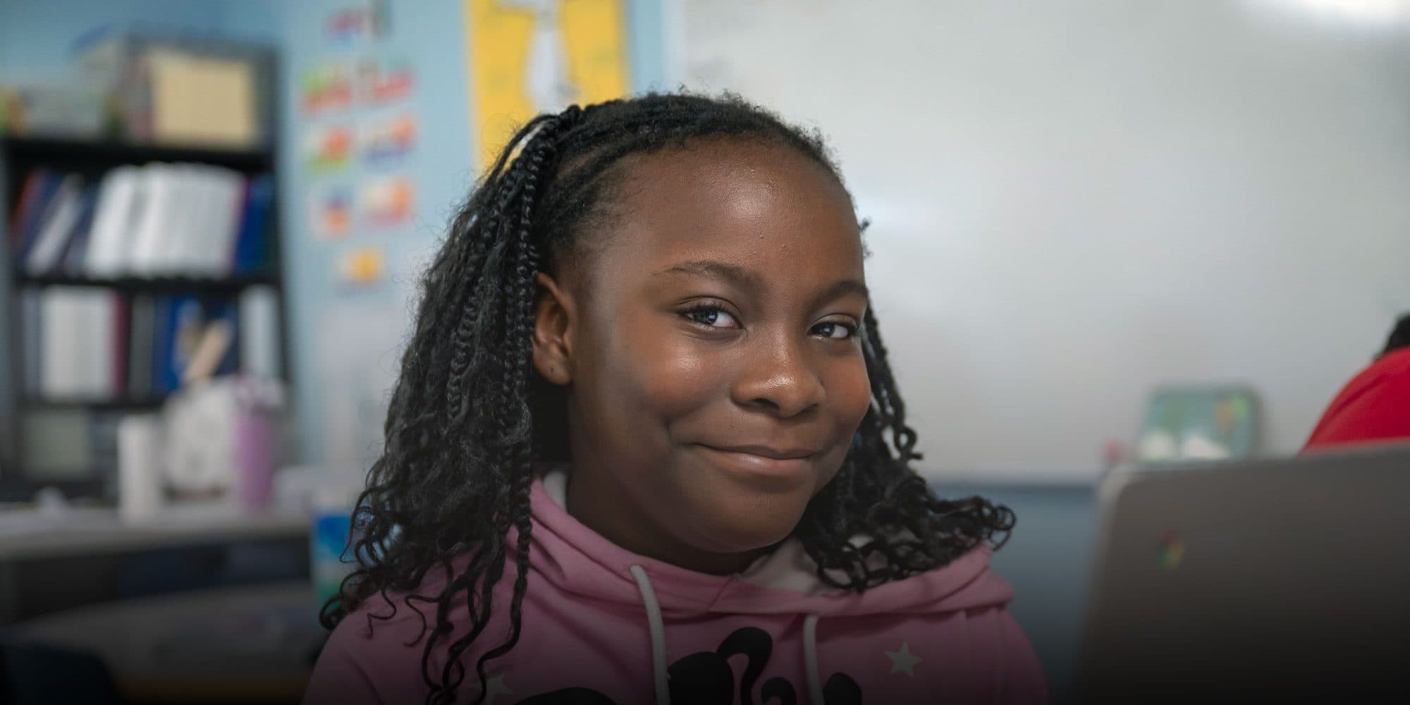 Smiling student with braids in a pink hoodie, sitting beside a laptop in a classroom.