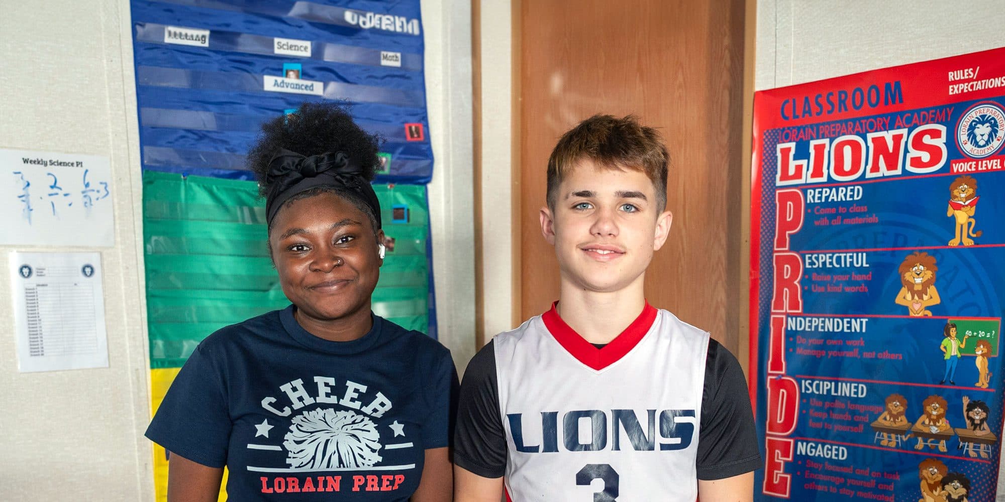 Two people in Lorain Prep cheer and Lions shirts smiling beside a Lions PRIDE classroom poster.