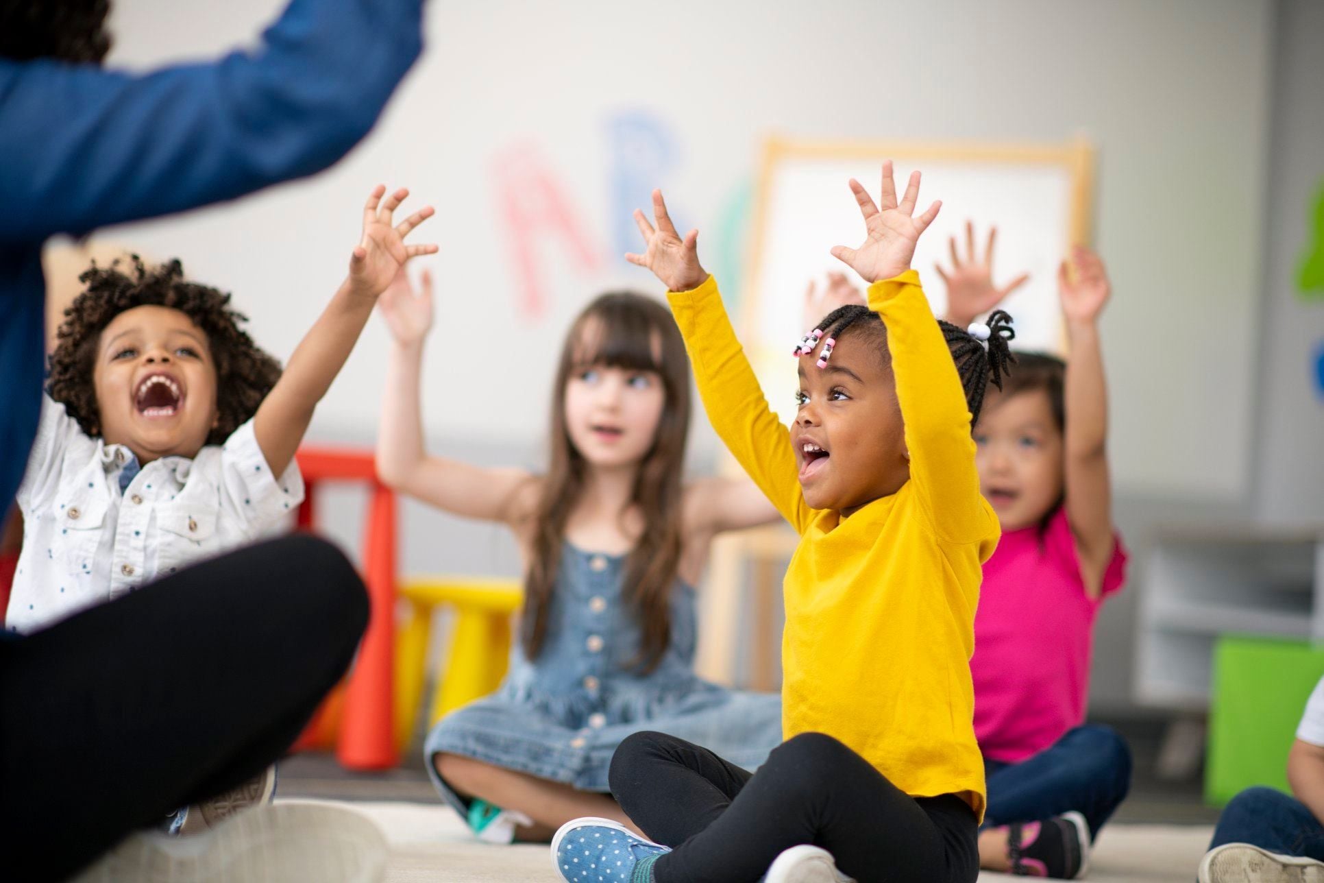Young children sitting on floor with arms raised, looking up.