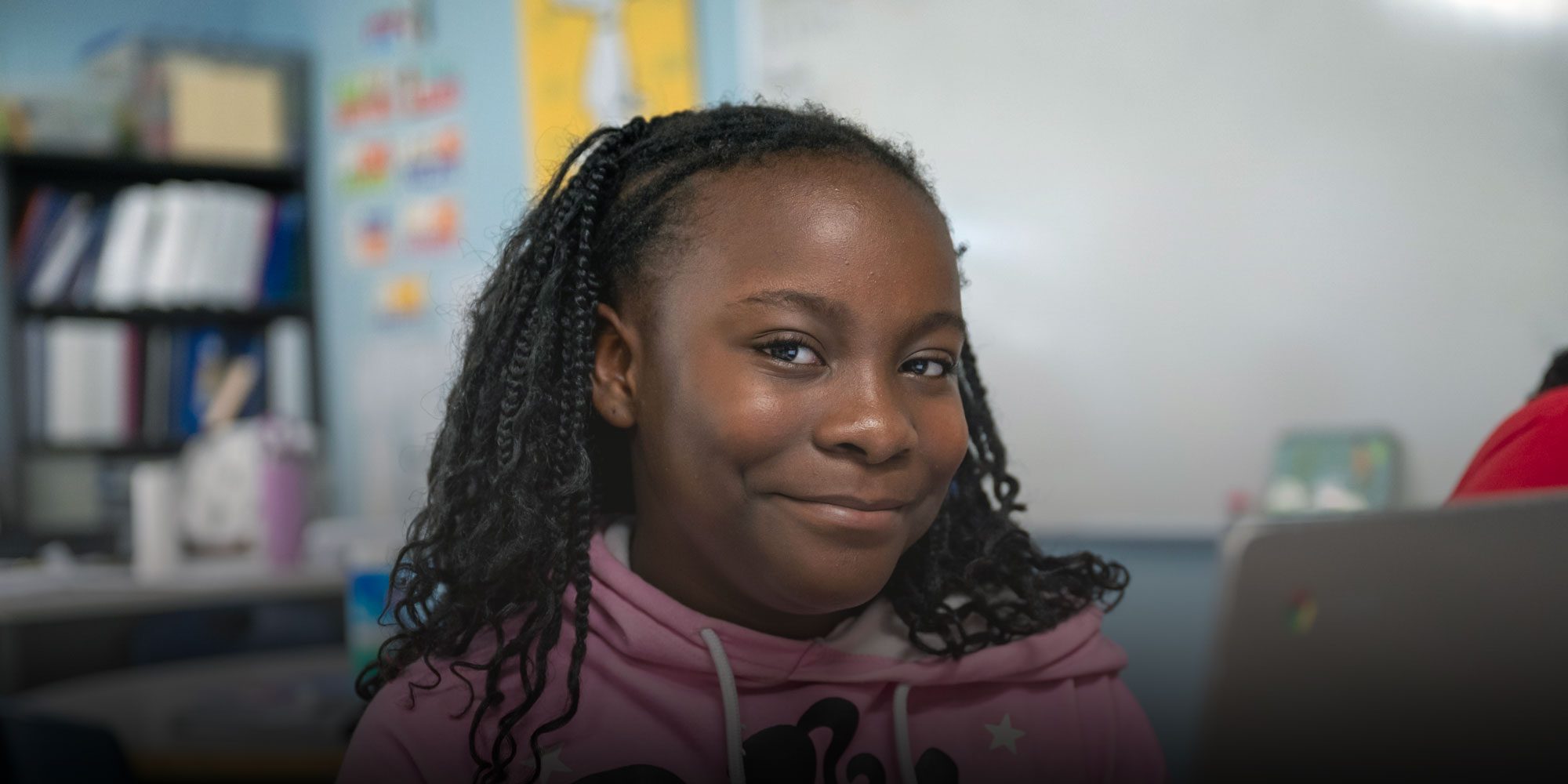 Smiling student with braids in a pink hoodie, sitting beside a laptop in a classroom.