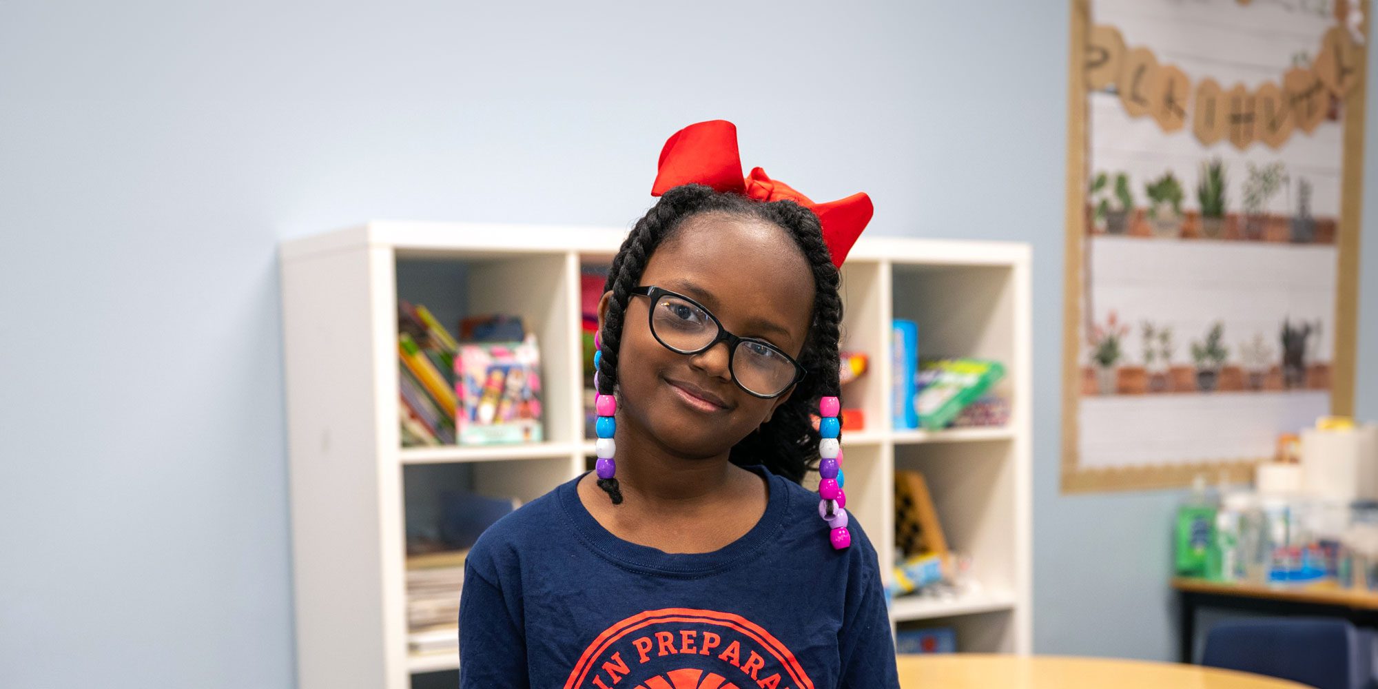 Smiling student wearing glasses, a red bow and a Lorain Prep shirt in a classroom.